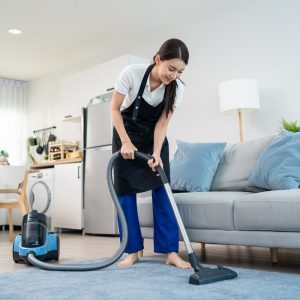 Asian cleaning service woman worker cleaning in living room at home. Beautiful young girl housekeeper cleaner wear apron and vacuuming messy dirty floor for housekeeping housework and chores in house.
