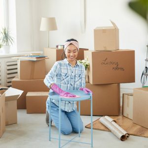 Full length portrait of cheerful Asian woman cleaning furniture while moving into new house with boxes in background, copy space